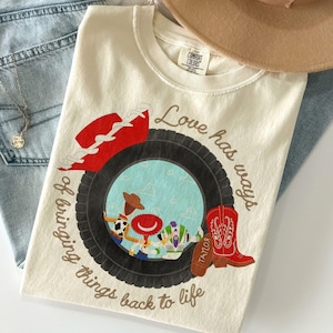 May include: A cream-colored t-shirt featuring a graphic of a black tire framing three cartoon characters. The text reads Love has ways of bringing things back to life. A red cowboy hat and a red cowboy boot with the name Taylor are also printed.