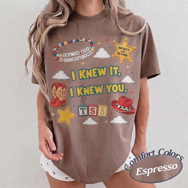 May include: A brown short-sleeve t-shirt featuring a graphic print with the text "I KNEW IT, I KNEW YOU," "TS5," "INFINITY AND BEYOND," and "You've got a friend in me." The design includes illustrations of a beaded bracelet, a sheriff star, cowboy boots, a hat, and clouds.