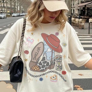 Puede incluir: Una camiseta color crema con un estampado gr&aacute;fico de una bota vaquera con patr&oacute;n de manchas de vaca, un sombrero rojo, el nombre Taylor, el n&uacute;mero 13 y bloques con las letras T y S. El dise&ntilde;o incluye peque&ntilde;as flores y estrellas.