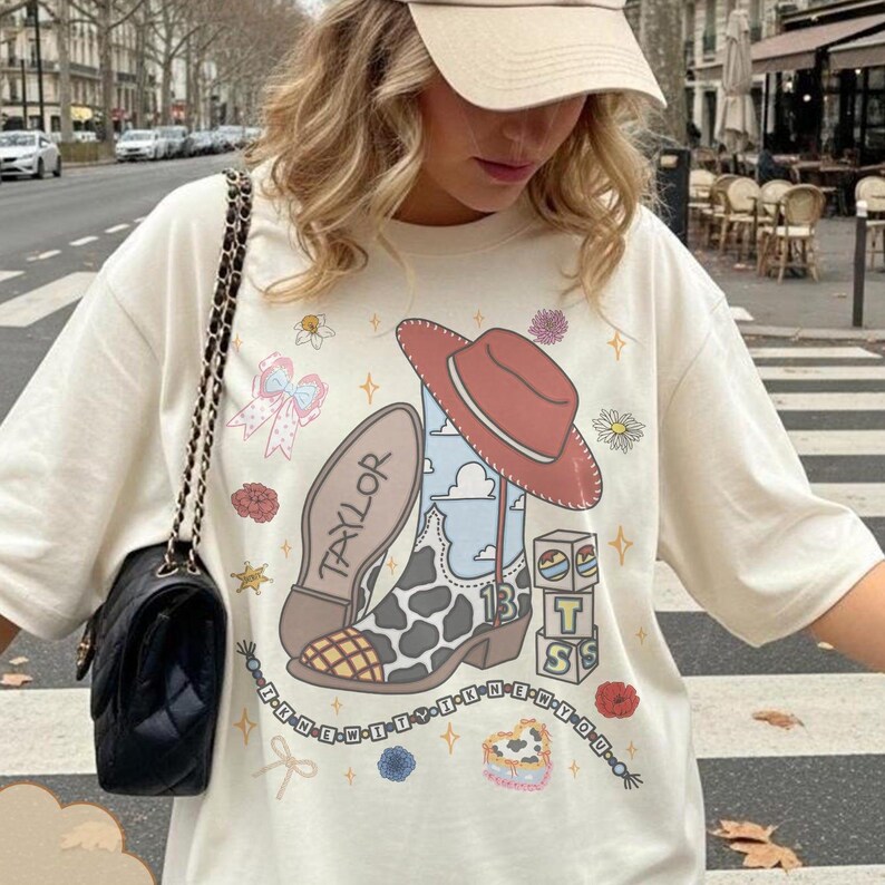 Puede incluir: Una camiseta color crema con un estampado gr&aacute;fico de una bota vaquera con patr&oacute;n de manchas de vaca, un sombrero rojo, el nombre Taylor, el n&uacute;mero 13 y bloques con las letras T y S. El dise&ntilde;o incluye peque&ntilde;as flores y estrellas.