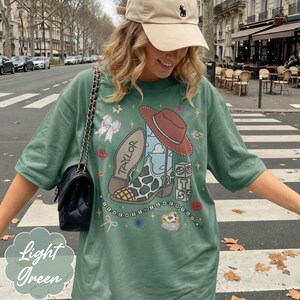 Puede incluir: Una camiseta verde claro con un estampado gr&aacute;fico de una bota de vaquero con manchas de vaca, un sombrero rojo, bloques con las letras T, S y el n&uacute;mero 13, un lazo y el nombre Taylor. Se combina con un bolso de hombro negro acolchado.