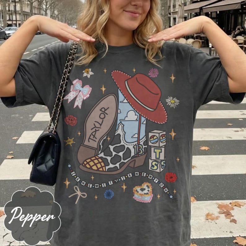 Puede incluir: Una camiseta gris carb&oacute;n con un estampado gr&aacute;fico de una bota de vaquero con estampado de vaca con el nombre Taylor, un sombrero de vaquero rojo, bloques con las letras T, S y el n&uacute;mero 13, un lazo y el texto I KNEW IT I KNEW YOU en un patr&oacute;n circular.