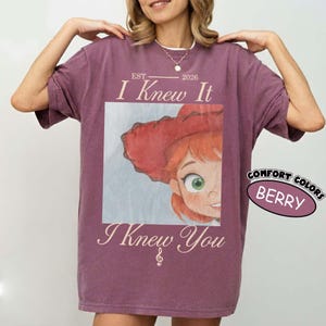 May include: A berry-colored short-sleeve t-shirt featuring a graphic print of a cartoon character with red hair and a red hat. The text reads EST 2026, I Knew It, and I Knew You, with a small musical note symbol. A sticker label reads COMFORT COLORS BERRY.