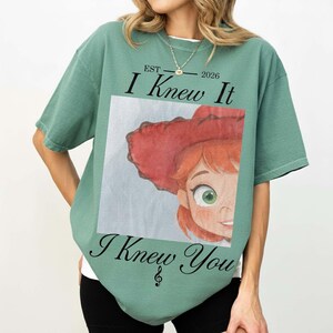 May include: A sage green short-sleeve t-shirt featuring a graphic print of a cartoon character with red hair and a red hat. The text reads EST 2026 I Knew It I Knew You with a treble clef symbol below the text. The design is centered on the chest.
