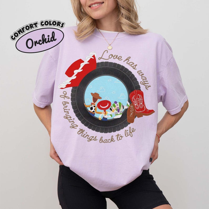 May include: An orchid-colored t-shirt featuring a graphic of a black tire framing cartoon characters. The design includes a red cowboy hat, a red cowboy boot with the name Taylor, and the text Love has ways of bringing things back to life.