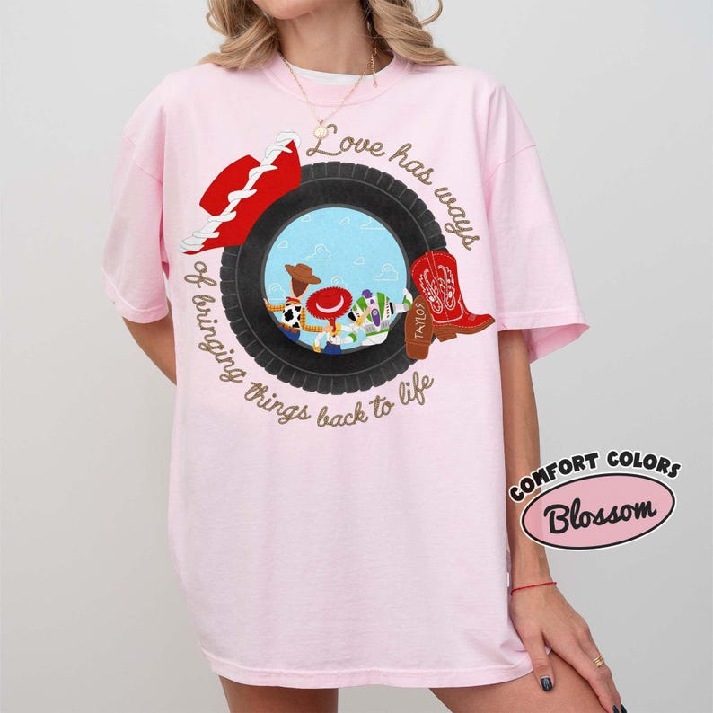 May include: A light pink t-shirt featuring a graphic of a black tire framing a blue sky scene with cartoon characters. The design includes a red hat, a red cowboy boot, and the text Love has ways of bringing things back to life in brown script.