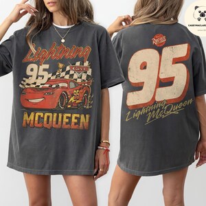 May include: A charcoal gray t-shirt featuring a graphic of the red race car Lightning McQueen with the number 95, a checkered flag, and the text Lightning McQueen on the front, and a large 95 with the text Lightning McQueen on the back.