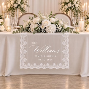 May include: A white lace table runner featuring the printed text THE Williams JAMES & SOPHIA 08.12.2026. The runner has intricate floral lace borders and is draped over a solid cream-colored tablecloth, topped with a large white floral centerpiece.