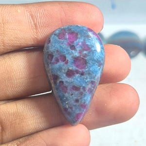 Peut inclure: Une pierre précieuse polie en forme de goutte d'eau présentant un motif tacheté d'inclusions rose magenta vif incrustées dans une matrice bleu ciel opaque et lisse. La pierre a une surface brillante et une taille cabochon symétrique.
