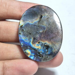 Könnte beinhalten: Ein ovaler Labradorit-Edelstein mit einer polierten, glatten Oberfläche, der irisierende Blitze in Blau, Gold und Violett auf einer dunkelgrauen Basis zeigt, durchzogen von natürlichen inneren Rissen und mineralischen Einschlüssen.