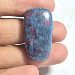May include: A polished, rectangular gemstone with rounded corners, featuring a mottled blue and grey base color interspersed with distinct, irregular magenta and deep pink inclusions throughout the surface of the stone.