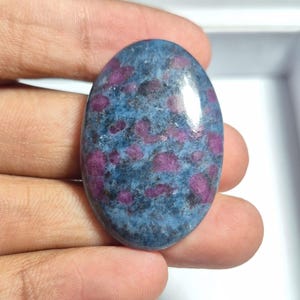Op de afbeelding: Een ovaalvormige cabochon edelsteen met een gepolijst oppervlak met een gevlekt patroon van diepblauwe en levendige magenta-roze insluitingen. De steen heeft een gladde, afgeronde bovenkant met een glanzende afwerking en natuurlijke kleuren.