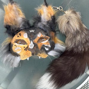 May include: A handmade, furry mask and tail set. The mask features orange, black, and white fur with a painted face design. The tail is a mix of brown, gray, and white fur, attached with a gold-colored clasp. The set is designed for cosplay or costume use.