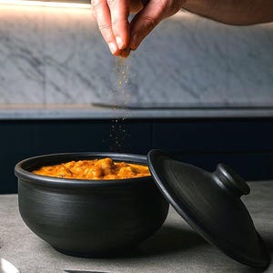 Puede incluir: Un cuenco de cerámica negro lleno de un plato cremoso anaranjado, con la tapa ligeramente abierta. Una mano espolvorea especias sobre la comida. Una cuchara, un cuchillo y hierbas están en la encimera.