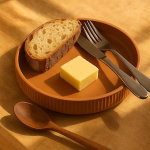 以下が含まれることがあります： 素朴なテラコッタの皿にパンのスライス、バターの正方形、フォーク、ナイフが乗っています。木製のスプーンが木製の表面に置かれています。温かい光が、シンプルで魅力的な食事を連想させます。