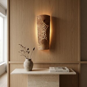 May include: A semi-cylindrical wall sconce made of textured, weathered brown clay featuring a vertical pattern of small, circular cutouts that emit a warm, soft glow. The light fixture is mounted on a vertical wood-paneled wall surface.