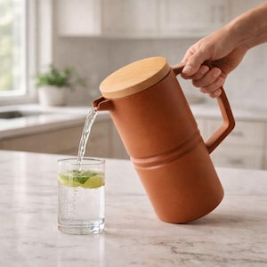 Puede incluir: Una jarra de color terracota con tapa de madera vierte agua en un vaso con rodajas de lima. La jarra tiene un asa y un pico. El vaso está sobre una encimera de mármol.