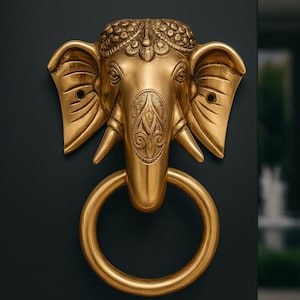 May include: A gold-colored elephant head door knocker with a large ring handle. The elephant has detailed carvings on its head and trunk, and is mounted on a dark surface. The knocker is a decorative piece.