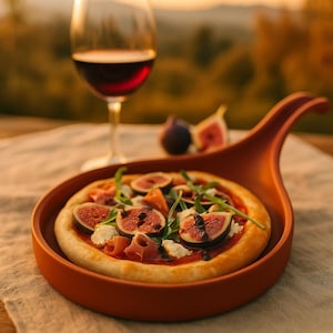 以下が含まれることがあります： テラコッタ色のサービング皿に、イチジク、プロシュート、ルッコラを乗せた小さなピザ。背景には赤ワインのグラスとイチジク。ピザは明るい色のテーブルクロスの上にあります。