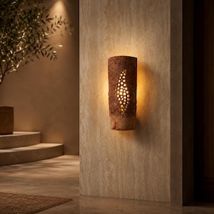 May include: A cylindrical wall sconce with a textured, brown, stone-like exterior featuring a vertical, leaf-shaped cutout pattern. The fixture emits a warm, golden light through the perforations, casting a soft glow against the wall surface.