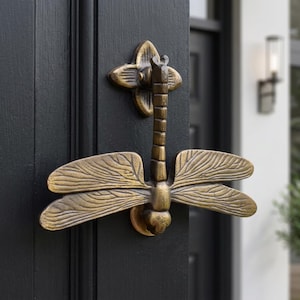 May include: A brass-toned metal dragonfly door knocker featuring a detailed segmented body and four wide, textured wings. The insect-shaped hardware is mounted on a black door, with a decorative four-petaled base plate at the top and a circular strike plate at the bottom.