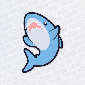 May include: A light blue cartoon shark sticker with a white belly and a pink mouth. The shark has black outlines and is smiling. The sticker is on a white background with a repeating pattern of the text "SIOBHAN MAKES".