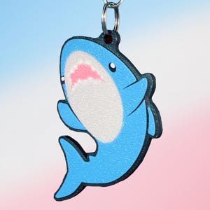 May include: A blue shark keychain featuring a white belly and a pink mouth. The shark is depicted in a cartoon style with a dark outline and is attached to a silver metal ring. The surface of the shark has a textured appearance and a glossy finish.