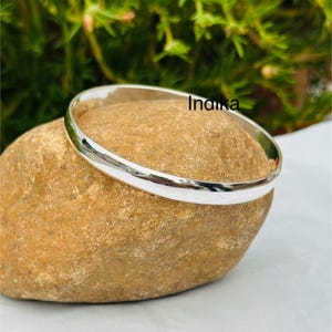 Pode incluir: Uma pulseira de bracelete prateada brilhante. A pulseira tem um design liso e arredondado, refletindo a luz. A palavra "Indika" está impressa na pulseira. A pulseira está sobre uma pedra castanha clara, com folhagem verde ao fundo.