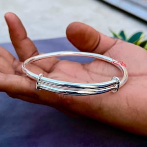 May include: A silver adjustable bangle. The bracelet features a straightforward design with two parallel bands and a sliding mechanism for size adjustment. The word "indika" is written in red script on the bracelet.