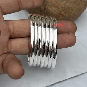 May include: A collection of polished silver bangles. The bangles are stacked together, reflecting light. The jewellery is shown on a hand, highlighting the smooth, rounded design and the gleam of the metal.