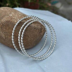 Op de afbeelding: Drie zilverkleurige armbanden met een gedraaid touwontwerp. De cirkelvormige armbanden liggen op een bruine steen. De sieraden worden gepresenteerd op een lichtblauwe ondergrond.