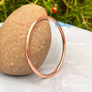 May include: A polished copper-colored bangle bracelet. The circular bracelet is displayed against a smooth, brown stone. The word "Balaji" is written in blue script on the right side of the image.