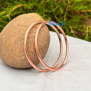 May include: Two rose gold bangles are displayed, resting on a white surface next to a large, brown stone. The bangles are circular and have a smooth, polished finish. The background features green foliage and the text "Balaji".
