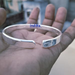 Puede incluir: Un brazalete de plata pulida con un diseño rectangular. El brazalete abierto presenta la palabra "indika" en texto azul. El brazalete se muestra en una mano.