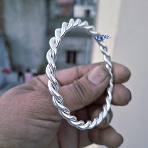 May include: A silver-colored, twisted metal bangle bracelet. The bracelet is held in a hand, showcasing its braided design. The bracelet's design is a series of interwoven strands, creating a textured, rope-like appearance. The bracelet is a piece of jewelry.