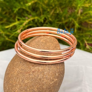 May include: Four copper-colored bangles stacked on a brown stone. The bangles are smooth and shiny, reflecting light. The background is a blurred mix of green and white. The word "Balaji" is written in blue.