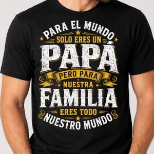 May include: A black short-sleeved t-shirt featuring white and yellow distressed-style text that reads: PARA EL MUNDO SOLO ERES UN PAP&Aacute; PERO PARA NUESTRA FAMILIA ERES TODO NUESTRO MUNDO, accented with small stars and decorative ribbon graphics.