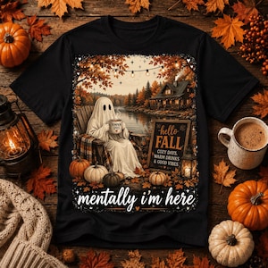 Może przedstawiać: Czarny t-shirt z grafiką ducha siedzącego na krześle nad jeziorem, trzymającego kubek z napisem Pumpkin Spice. Wzór zawiera chatę, dynie, lampki i znak z napisem Hello Fall, Cozy Days, Warm Drinks & Good Vibes, z tekstem Mentally I'm Here u dołu.
