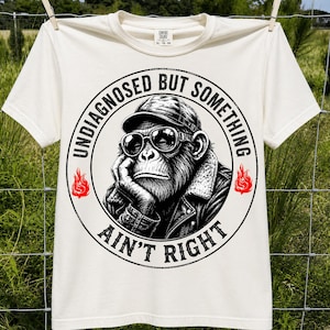 May include: An off-white short-sleeve t-shirt featuring a circular graphic of a chimpanzee wearing a cap, sunglasses, and a jacket. The text "UNDIAGNOSED BUT SOMETHING AIN'T RIGHT" surrounds the monkey, with two small red flame icons on the sides.