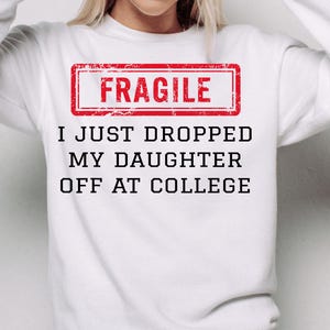 Könnte beinhalten: Weißes Sweatshirt mit dem Text "FRAGILE" in einem roten Rechteck und dem Satz "I JUST DROPPED MY DAUGHTER OFF AT COLLEGE" in Schwarz. Ein lässiger Stil.