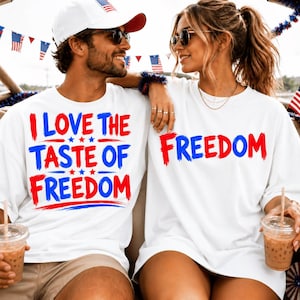 Op de afbeelding: Twee witte T-shirts met korte mouwen en rode en blauwe grafische tekst. Het linker T-shirt toont de tekst I LOVE THE TASTE OF FREEDOM met kleine sterretjes. Het rechter T-shirt toont de tekst FREEDOM in een vetgedrukt penseelstijl lettertype.