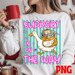 May include: White sweatshirt with a graphic featuring a cartoon duck carrying a bag filled with school supplies. The text "SUPPORT IS ON THE WAY" is displayed in pink and teal. A red mug is held in the upper left corner.