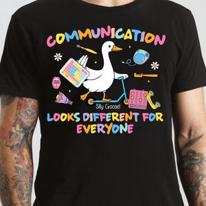 Puede incluir: Camiseta negra con un gráfico colorido que presenta un ganso blanco en un patinete azul, sosteniendo un teclado y un pincel. El texto dice "COMMUNICATION LOOKS DIFFERENT FOR EVERYONE" en colores arcoíris. El ganso está etiquetado como "Silly Goose!"