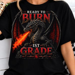 May include: A black t-shirt featuring a graphic of a black dragon with red wings breathing fire. The text reads "READY TO BURN" above the dragon and "1ST GRADE" below it in a distressed red font. The design includes a small circular emblem.
