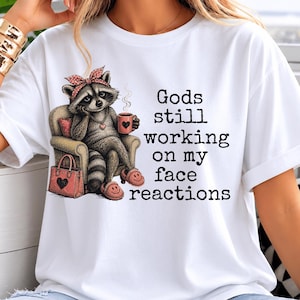 Può includere: Una t-shirt bianca con l'illustrazione di un procione seduto su una poltrona, che indossa una fascia a pois e pantofole, mentre tiene una tazza di caffè. Il testo recita: Gods still working on my face reactions. Una borsa rosa con un cuore.
