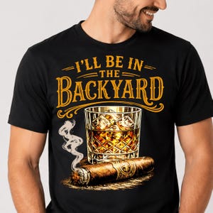 May include: A black t-shirt featuring a graphic print of a glass of amber liquid with ice cubes and a lit cigar with rising smoke. Above the graphic, the text reads "I'LL BE IN THE BACKYARD" in a bold, stylised yellow font with decorative lines.