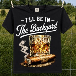 May include: A black short-sleeve t-shirt featuring the white text I'LL BE IN The Backyard above a graphic of a glass of amber liquid with ice cubes and a lit cigar with rising smoke. The shirt has a Comfort Colors brand label at the neck.
