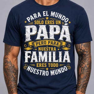 May include: A navy blue short-sleeve t-shirt featuring white and yellow distressed-style text that reads: PARA EL MUNDO SOLO ERES UN PAPA PERO PARA NUESTRA FAMILIA ERES TODO NUESTRO MUNDO, accented with small yellow stars and decorative banners.