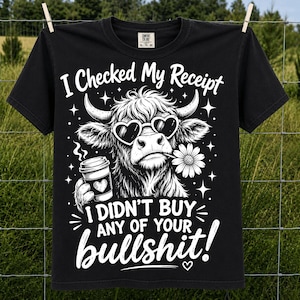 Może przedstawiać: Czarny t-shirt z białą grafiką przedstawiającą krowę rasy Highland w okularach przeciwsłonecznych w kształcie serc, trzymającą kubek kawy i stokrotkę. Napis na koszulce brzmi: I Checked My Receipt I Didn't Buy Any Of Your bullshit! Krótki rękaw.