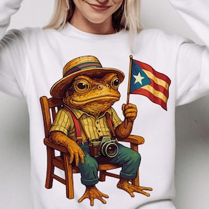 May include: A white sweatshirt featuring a cartoon frog wearing a straw hat, striped shirt, suspenders, and blue jeans. The frog is holding a Puerto Rican flag and a camera while sitting in a wooden chair.